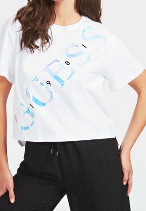 Vrouw draagt een wit cropped T-shirt met holografische tekst "GUESS 1981" en zwarte broek met subtiele verticale strepen, poseert met hand op heup.
