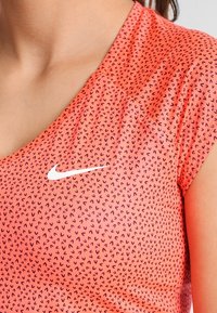 Nike sportstopp i ljus korall med ett subtilt mönster av svarta abstrakta former. Har en V-ringning och korta ärmar; ikonisk vit logotyp på bröstet.