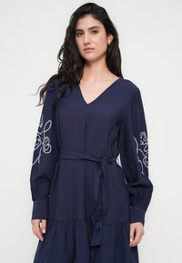 Μπλε φορεμα με V-λαιμόκοψη, μακριά μανίκια με λευκή κεντημένη ανθοκομική διακόσμηση, εφαρμοστή ζώνη στη μέση και φούστα με επίπεδα. Μαλακή υφή.