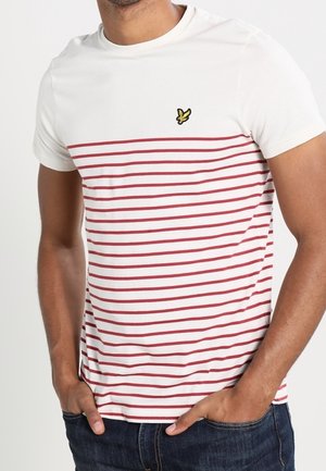 Mand iført hvid T-shirt med tynde røde horisontale striber og lille gul fuglelogo på brystet, kombineret med mørkeblå jeans.