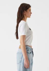Camiseta blanca corta con mangas cortas y texto negro en el pecho, combinada con unos jeans de tiro alto de color azul claro. Diseño sencillo.