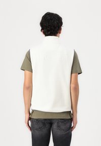 Weißer Fleece-Weste mit hohem Kragen, mit eingraviertem "HACKETT"-Logo auf dem Rücken, getragen über einem grünen Kurzarmhemd und dunklen Jeans.