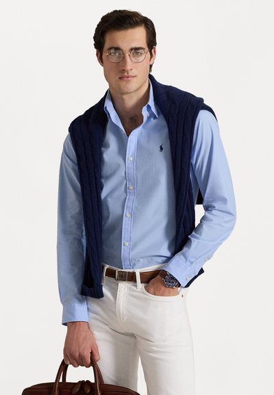 Gaiši zilā pogā krūšjaka ar tumši zilu adītu džemperi, kas uzmest uz pleciem, baltas bikses, brūns josta un brūna ādas soma.