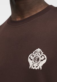 Κοντινό πλάνο ενός ατόμου που φοράει καφέ πουκάμισο με ένα λευκό κεντητό διακοσμητικό λογότυπο που απεικονίζει ένα στιλιζαρισμένο γράμμα «C» στο στήθος.