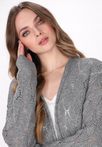 Šedý pletený cardigan s vzorem listů, otevřený design a dlouhé rukávy. Nosený přes bílý top, který má jemný, texturovaný povrch.