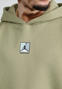 Olivengrøn hoodie lavet af blødt bomuld med en kængurulomme, der har et Jordan-logo i sort og blå på brystområdet.