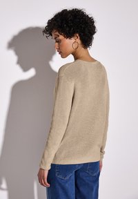 Béžový pletený svetr s texturovaným povrchem, dlouhými rukávy a kulatým výstřihem, doplněný modrými džíny. Stín viditelný na zdi.