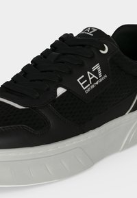 Μαύρο sneaker EA7 Emporio Armani με μαύρα κορδόνια, δίχτυ στα πλαϊνά και χοντρή λευκή σόλα σε ανοιχτό φόντο.