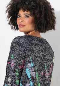 T-shirt à manches longues en noir avec un motif texturé, présentant des éclaboussures vibrantes de couleur rose, bleue et verte sur le tissu.