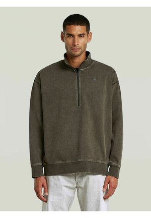 Mann trägt einen dunkelgrauen Quarter-Zip-Sweatshirt mit Nahtdetails und hellgraue Hose, steht vor einem schlichten hellen Hintergrund.