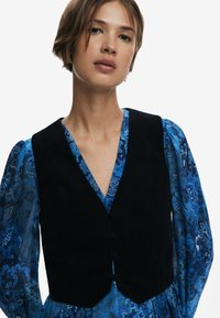 Crni kordurov gilet s V-izrezom, prilagođenim krojem i suptilnim kopčanjem, složen preko plave bluze s uzorkom.