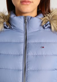 Femme portant une doudoune bleu clair avec une capuche bordée de fourrure et une fermeture éclair frontale, arborant un petit logo rouge, blanc et bleu sur la poitrine.