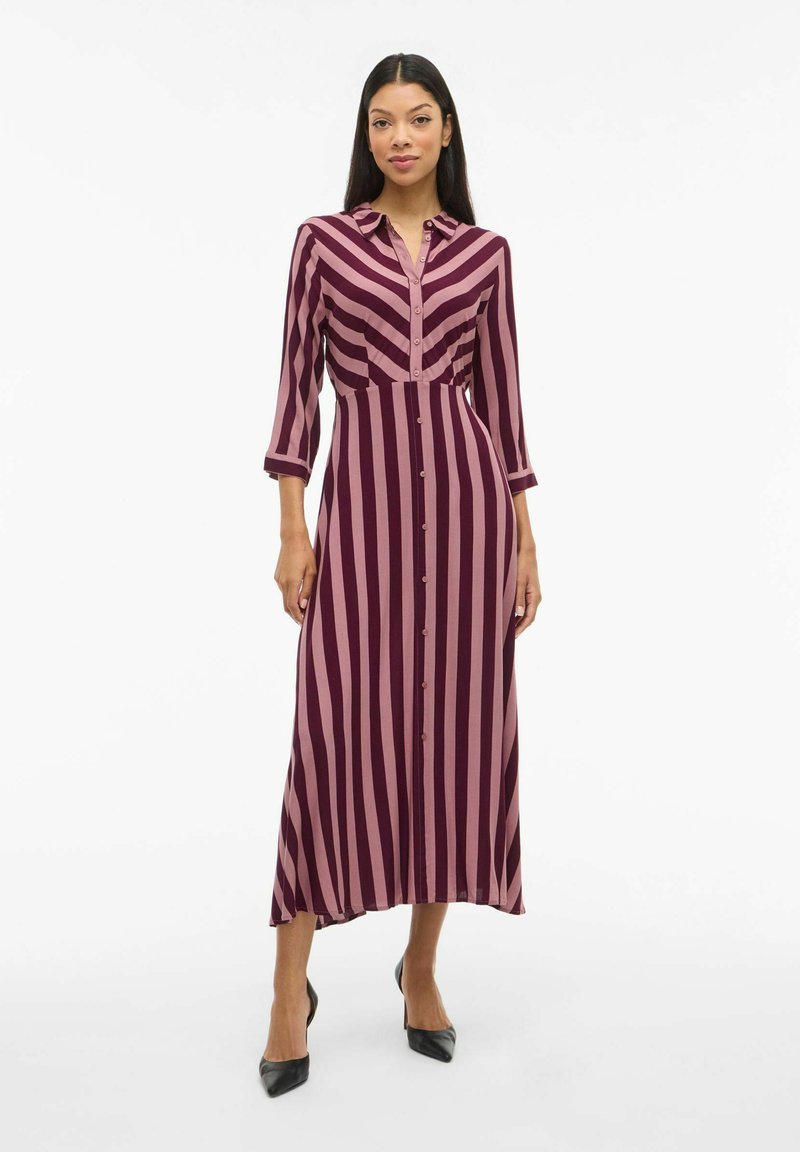 Langes Hemdblusenkleid mit kastanienbraunen und rosa vertikalen Streifen, ausgestattet mit einem Kragen, Dreiviertelärmeln und einem geraden Saum. Getragen mit schwarzen High Heels.