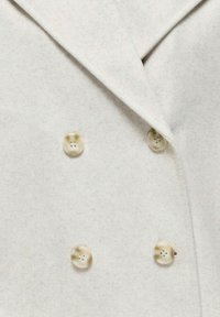 Gros plan d'un manteau en laine beige montrant quatre boutons brun clair disposés en style croisé sous un col à large revers.