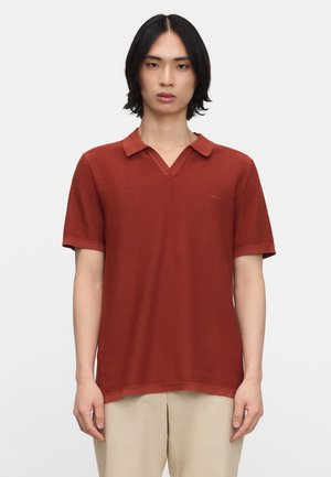 Jong persoon met schouderlang zwart haar, gekleed in een roestrood poloshirt met korte mouwen en beige broek tegen een egale achtergrond.