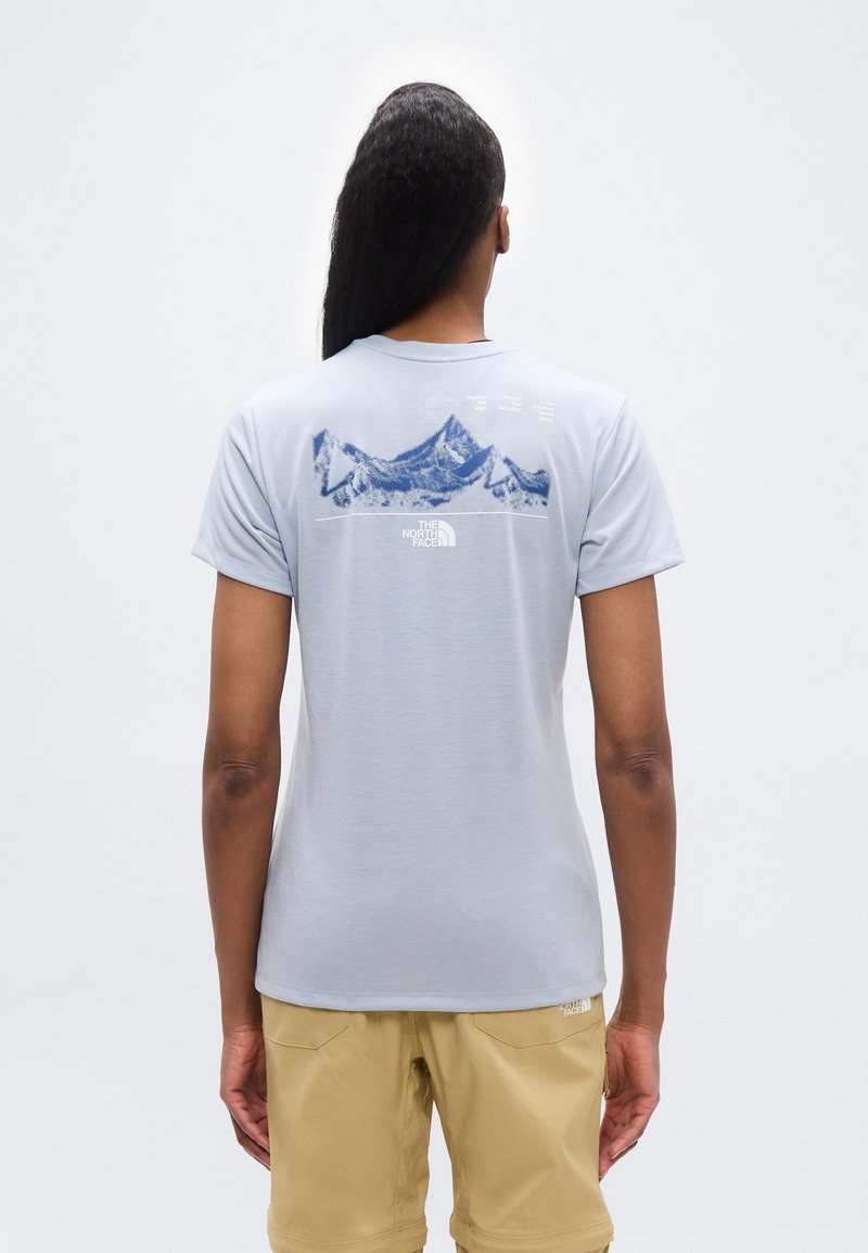 Άτομο με μακριά σκούρα μαλλιά, που φοράει ανοιχτό μπλε μπλουζάκι με γραφικό βουνού και μπεζ σορτς, στέκεται με την πλάτη γυρισμένη σε απλό φόντο.