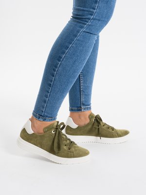 Person trägt blaue Skinny-Jeans und olivgrüne Wildledersneaker mit weißen Sohlen, stehend auf weißem Hintergrund.