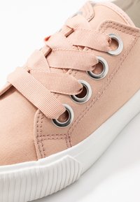 Zbliżenie różowego płóciennego trampka z szerokimi różowymi sznurówkami przeplecionymi przez cztery metalowe oczka oraz białą gumową podeszwą.