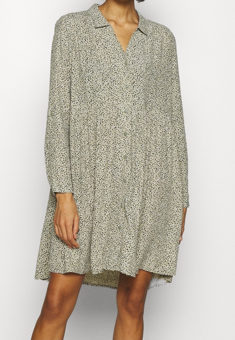 Femme portant une robe ample à manches longues boutonnée avec un petit motif à pois noir et blanc et un col, debout devant un fond uni.