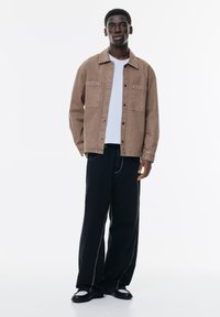 Kahvin denim-takki, jossa on kaksi rintataskua, yhdistetty valkoiseen t-paitaan ja hulmuaviin mustiin housuihin, joissa on valkoista ommelta. Sujuva pinta.