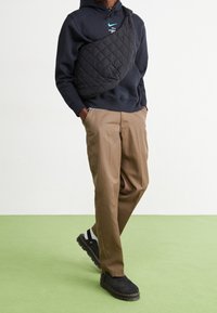 Svart quiltad axelväska bärs över en mörk hoodie, tillsammans med khakifärgade byxor och svarta skor, mot en ljus bakgrund och grön golv.