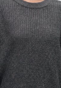 Gros plan d'une personne portant un pull en maille gris foncé avec un col rond côtelé.
