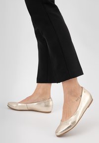 Ballerines métalliques dorées avec une texture lisse, un bout arrondi et un profil bas, associées à un pantalon noir cropped.