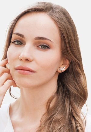 Silberne Libellenohrringe mit filigranen Details, getragen von einer Person mit langen, glatten Haaren. Einfache weiße Kleidung im Hintergrund.