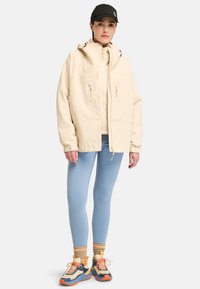 Beige softshell jacket with a high collar and two zippered pockets, worn over light blue leggings and colorful sneakers. Black cap on head.