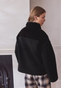 Veste en polaire noire avec un col montant et une texture douce, présentant un dos uni et des détails en tissu contrastant. Associée à un pantalon à carreaux.