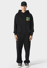 Sudadera negra con un patrón colorido de hojas en el bolsillo del pecho, combinada con pantalones de chándal negros y zapatillas negras. Una gorra sencilla completa el conjunto.