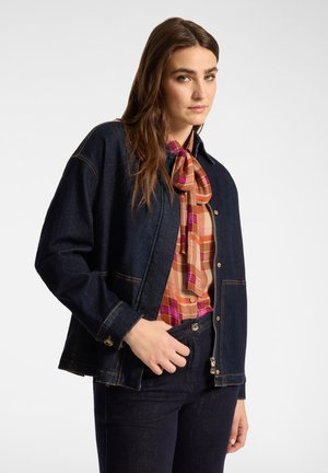 Femme aux longs cheveux bruns portant une veste en jean foncé et un pantalon au-dessus d'une chemise à carreaux colorée avec un col lavallière, debout devant un fond uni.