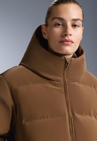 Smeđa opcija jakne s podstavom koja ima visoki ovratnik i skrivenu munju. Glatka tekstura s elegantnim, modernim dizajnom. Vidljiv detalj remena.