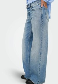 Jeans de mezclilla de corte ancho en azul claro con textura desvaída, bolsillos delanteros y corte recto, combinados con zapatos negros.