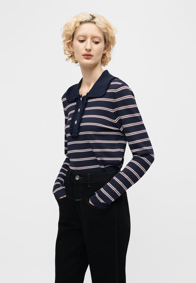 Νεαρή γυναίκα με κοντά, ξανθά, σγουρά μαλλιά, ντυμένη με μακρυμάνικη μπλούζα σε σκούρο μπλε με ρίγες και γιακά με βολάν, και μαύρο παντελόνι ψηλόμεσο, με τα χέρια στις τσέπες.