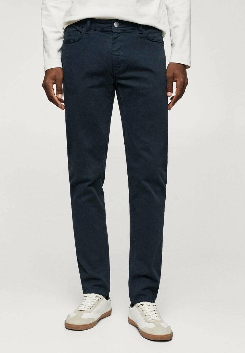 Homme portant un pantalon slim-fit bleu marine foncé et des baskets blanches, debout devant un fond clair uni.