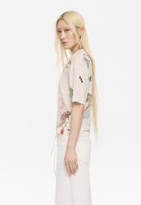 Top a maniche corte di un leggero beige con ricami floreali in diversi colori. Presenta una vestibilità comoda e una texture morbida. Indossato con pantaloni bianchi con coulisse.
