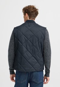 Mörk quiltad väst med hög krage, som har diagonal sömnad, lätt tyg och en slät yta, som är lager på lager över en grå tröja.