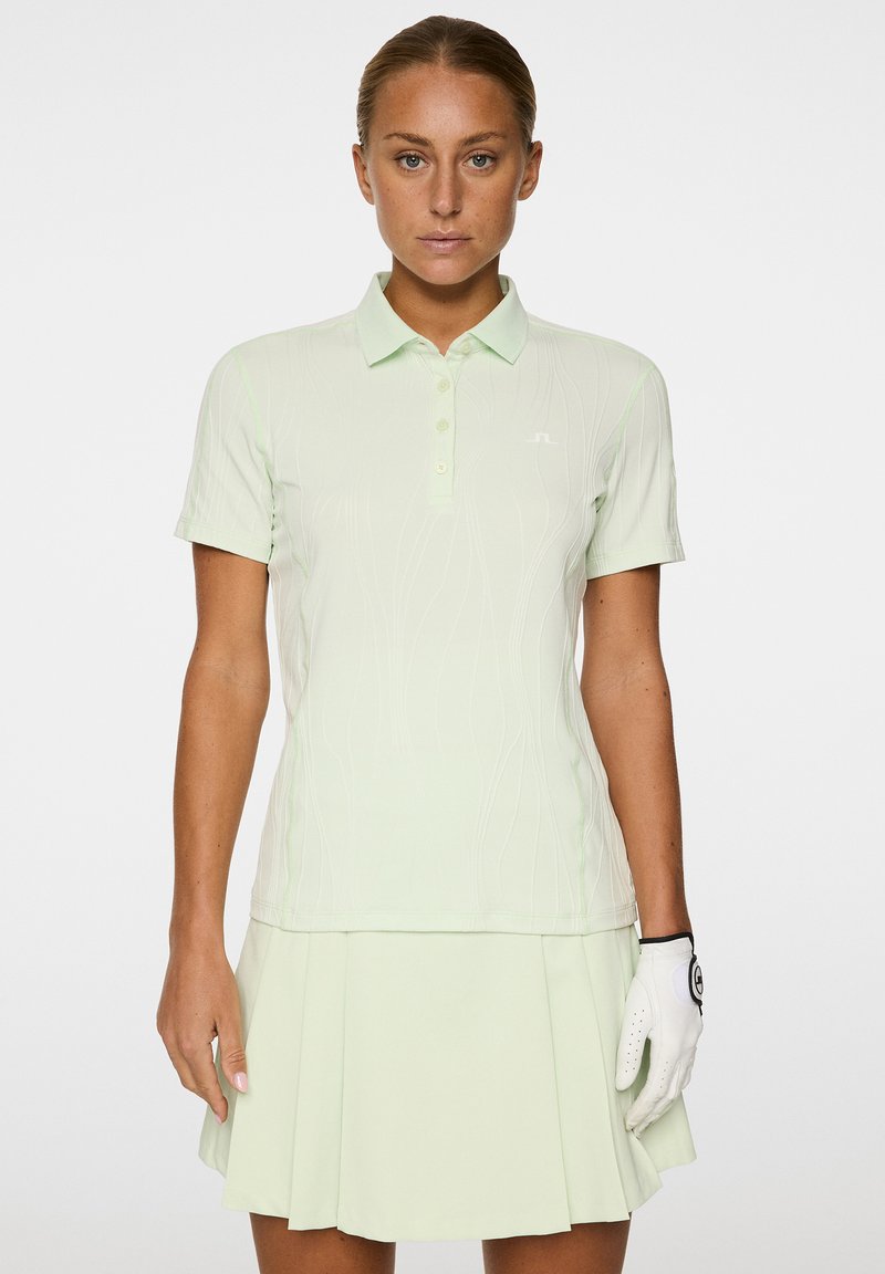 Hellgrünes Golf-Polo mit strukturiertem Muster und kurzen Ärmeln, ausgestattet mit einer Knopfleiste mit drei Knöpfen. Kombiniert mit einem passenden plissierten Rock.