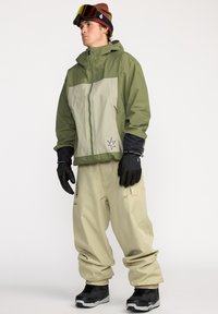 Kurtka snowboardowa w oliwkowym zielonym i beżowym kolorze, z kapturem, zamkiem błyskawicznym z przodu oraz detalem logo. Połączona z luźnymi beżowymi spodniami i czarnymi rękawiczkami.