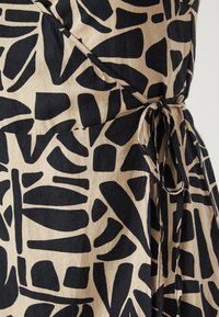 Motif abstrait noir sur tissu beige, avec un détail de nouage à la taille. La texture semble lisse et légère.