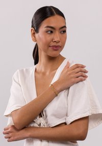 Goldene Kettenarmband, das an einem Handgelenk getragen wird und aus ineinander greifenden Gliedern besteht. Das Modell trägt ein leichter Leinenoberteil mit kurzen Ärmeln, das das Armband zur Schau stellt.