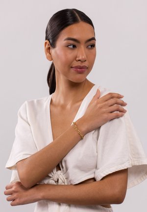 Gold chain bracelet worn on a wrist, featuring interlocking links. The model has a light linen top with short sleeves, showcasing the bracelet.