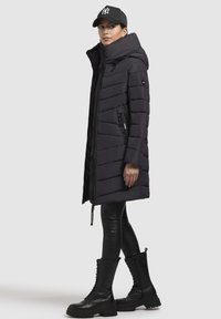 Doudoune noire avec un col montant et un motif matelassé diagonal, dotée de poches zippées sur les côtés. Associée à des leggings noirs et des bottes à lacets.