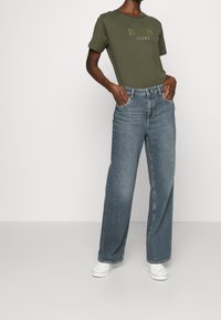 Personne portant un t-shirt MOSCH olive, un jean bleu taille haute large et des baskets blanches, les mains dans les poches, contre un fond blanc.