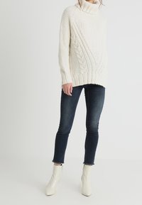 Gruba, kremowa sweter z warkoczowym splotem, luźny krój, w połączeniu z ciemnoniebieskimi skinny jeansami i białymi botkami na małym obcasie.