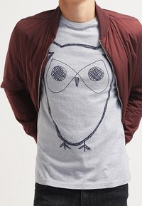 Chaqueta bomber burdeos sobre una camiseta gris que presenta un diseño de búho dibujado a mano. La tela parece suave y tiene un corte relajado.