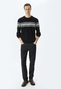 Svart stickad tröja med vita och grå horisontella ränder, som kombineras med mörka denimjeans och bruna ankelstövlar. Modellen poserar med händerna i fickorna.