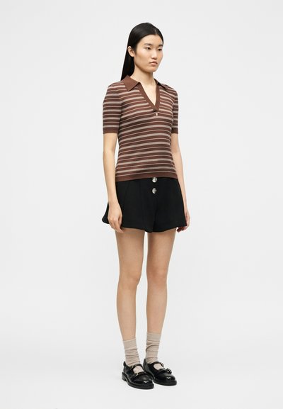 Femme debout portant un polo à manches courtes rayé marron, un short noir boutonné, des chaussettes beiges et des mocassins noirs sur un fond uni.