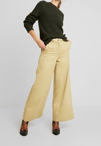Pantaloni beige a gamba larga in cotone, con tasche laterali e una vita alta, abbinati a un maglione verde scuro dalla texture.